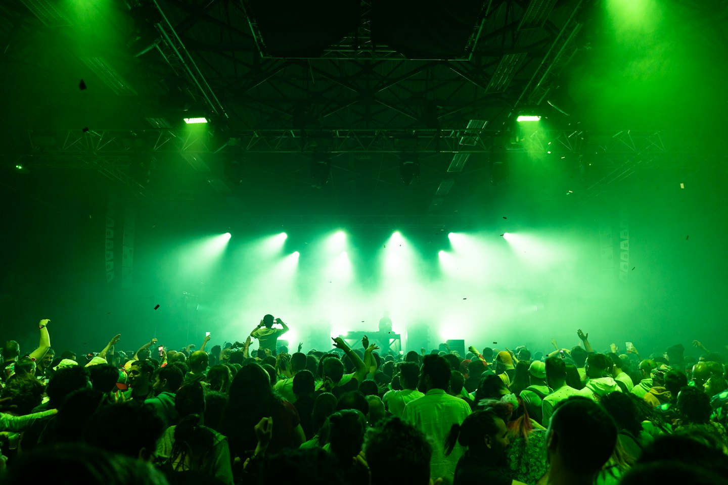 Crowd dancing with hands raised at a concert with green stage lights.